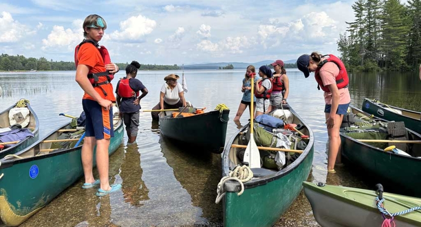 A group of people stand in ankle-deep water beside beached canoes. 